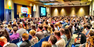 An audience in a conference setting, all facing a stage off-screen to the left.